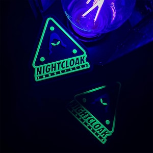May include: Two triangular patches with the text "NIGHTCLOAK INDUSTRIES" and an alien silhouette. The patches are black with neon green accents and glow in the dark. The image is taken in a dark setting with a purple light bulb in the background.