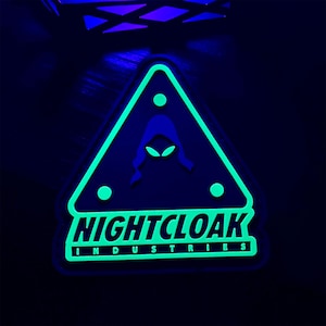 May include: A neon green and black patch with the text "NIGHTCLOAK INDUSTRIES". The patch features a triangular design with a hooded figure and three green dots. The design glows in the dark.