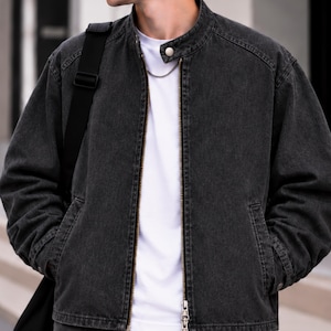 Könnte beinhalten: Eine anthrazitfarbene Jeans-Bomberjacke mit Stehkragen und einem Druckknopf, einem durchgehenden Metallreißverschluss und zwei Seitentaschen. Die Jacke wird über einem schlichten weißen T-Shirt getragen und hat einen schwarzen Schulterriemen.