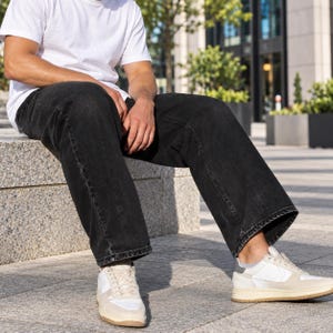 May include: A plain white short-sleeved t-shirt paired with loose-fitting black denim jeans. The outfit is completed with low-top sneakers featuring white panels, light beige suede overlays, and a dark grey heel tab, set on a thick rubber sole.