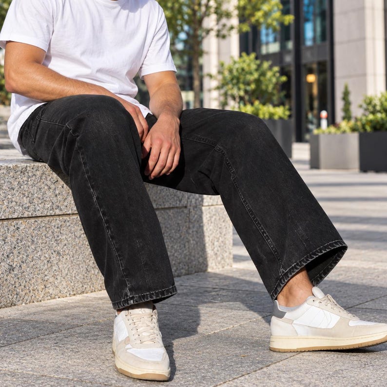 May include: A plain white short-sleeved t-shirt paired with loose-fitting black denim jeans. The outfit is completed with low-top sneakers featuring white panels, light beige suede overlays, and a dark grey heel tab, set on a thick rubber sole.