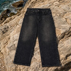 May include: A pair of high-waisted jeans in a faded black wash featuring a wide-leg silhouette, a classic five-pocket design, a button and zipper fly closure, and visible tonal stitching along the seams and pockets of the denim fabric.