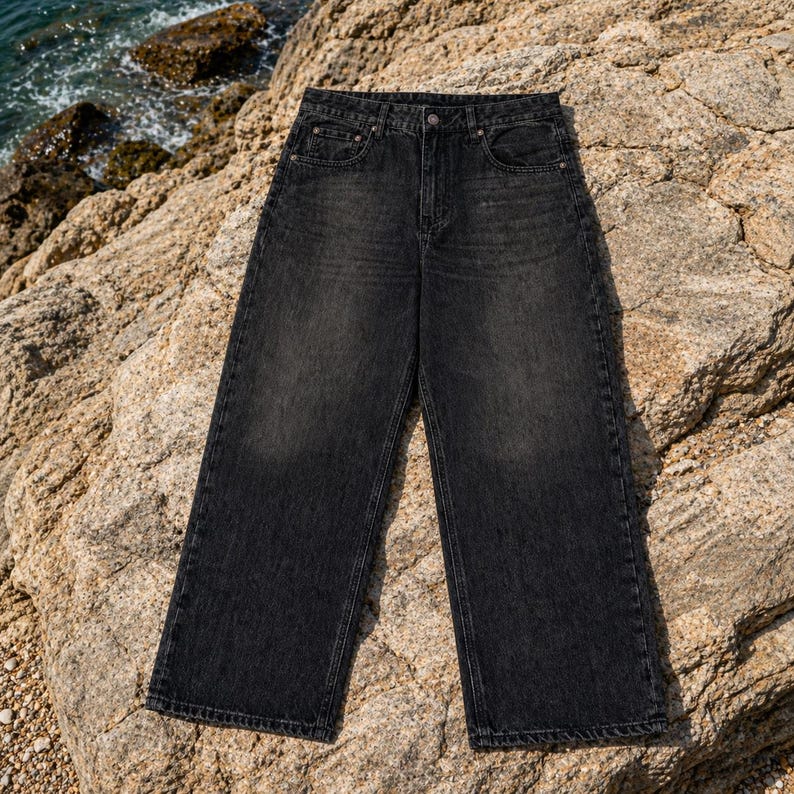 May include: A pair of high-waisted jeans in a faded black wash featuring a wide-leg silhouette, a classic five-pocket design, a button and zipper fly closure, and visible tonal stitching along the seams and pockets of the denim fabric.