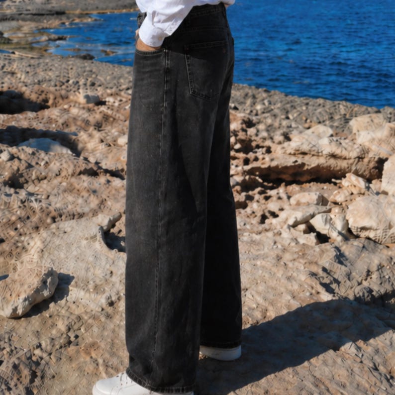 May include: A pair of loose-fitting, high-waisted black denim jeans with a straight-leg cut, featuring a classic five-pocket design, visible stitching along the seams, and a button fly closure, paired with a white long-sleeved shirt and white sneakers.
