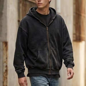 Puede incluir: Una sudadera negra con cremallera, capucha con cordón y bolsillo delantero. La sudadera tiene un aspecto vintage y lavado y una cremallera color latón. El modelo lleva debajo una camiseta negra y vaqueros azules.