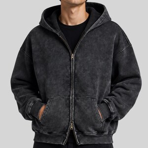 Puede incluir: Una sudadera con capucha y cremallera en color gris carbón con una textura de tela lavada y desgastada. Esta prenda incluye capucha, mangas largas con puños acanalados, una cremallera frontal completa y dos bolsillos tipo canguro. Se lleva sobre una camiseta negra.