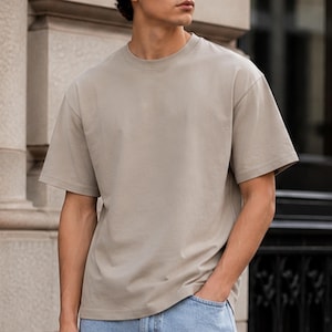 Könnte beinhalten: Beiges T-Shirt mit Rundhalsausschnitt und kurzen Ärmeln. Das Shirt besteht aus einem weichen Material und hat eine lockere Passform. Das Model trägt eine blaue Jeans. Das Shirt hat eine neutrale Farbe und kann mit vielen verschiedenen Outfits getragen werden.