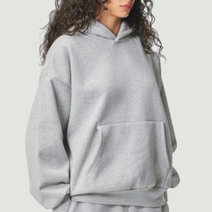 Puede incluir: Una sudadera con capucha gris claro con un bolsillo de canguro. La sudadera tiene una capucha con cordón y mangas largas. La tela parece ser un material suave y cómodo. El estilo general es casual y relajado.