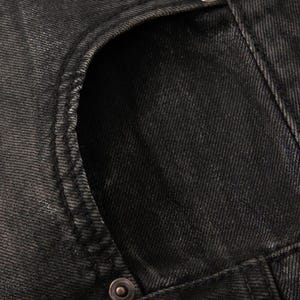 May include: A close-up view of black denim fabric featuring a curved pocket opening with visible stitching along the edge. A single metallic rivet is positioned at the base of the pocket seam, highlighting the texture of the dark cotton twill material.