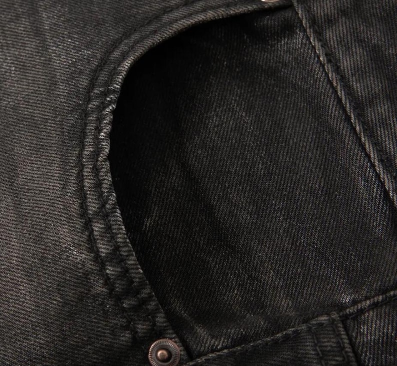 May include: A close-up view of black denim fabric featuring a curved pocket opening with visible stitching along the edge. A single metallic rivet is positioned at the base of the pocket seam, highlighting the texture of the dark cotton twill material.