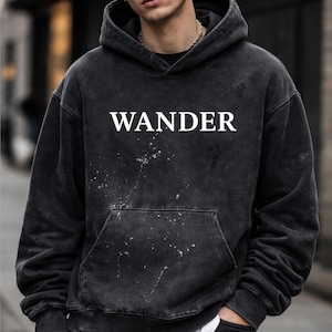 May include: A black hooded sweatshirt with the word "WANDER" in white block letters across the chest. The sweatshirt has a distressed look with white paint splatters and a front pocket.