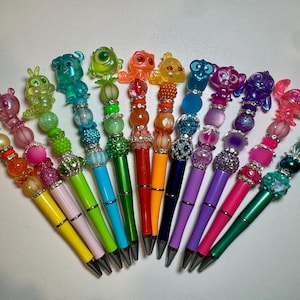 May include: A collection of colorful pens with decorative tops. Each pen features a different character figurine and a beaded design in various colors, including pink, green, blue, and orange. The pens have silver accents.