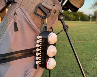 May include: A black golf ball and tee holder attached to a grey golf bag. The holder contains three golf balls and several white tees. The golf bag is on a golf course with clubs visible in the background.