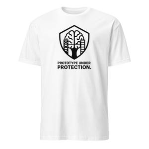 May include: White t-shirt featuring a black graphic of a brain inside a shield, with the text "PROTOTYPE UNDER PROTECTION." The design includes architectural elements and a tree-like structure, creating a unique and symbolic image.