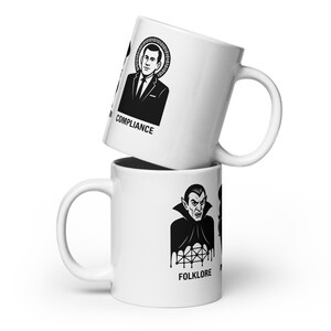 May include: Two white ceramic mugs stacked on top of each other. The top mug features a black and white illustration of a man with the word "COMPLIANCE" below. The bottom mug has a black and white illustration of a vampire with the word "FOLKLORE" below.