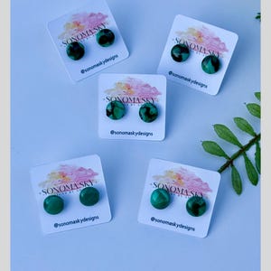 May include: Five pairs of round, green and black earrings on white cards. Each card has the text "SONOMA SKY" and "@sonomaskydesigns". The earrings are displayed on a light purple background with green leaves.