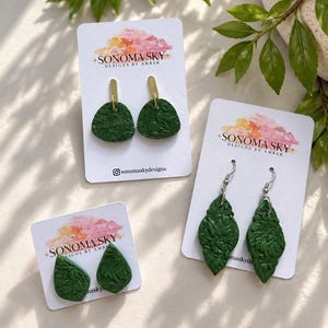 May include: Three pairs of dark green textured earrings displayed on white Sonoma Sky Designs by Amber cards. One pair features gold-tone posts with semi-circular pendants, another has elongated diamond shapes, and the third has teardrop shapes.