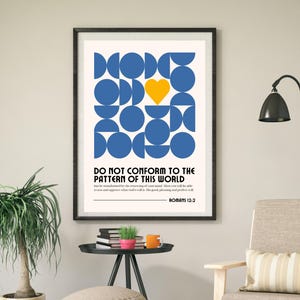 Op de afbeelding: Een ingelijste kunstdruk met een geometrisch ontwerp van blauwe cirkels en een gele hart. De tekst "DO NOT CONFORM TO THE PATTERN OF THIS WORLD" staat eronder, met een bijbelversverwijzing. Het kunstwerk zit in een zwarte lijst.
