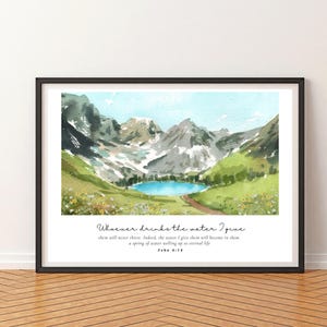 May include: Framed watercolour print depicting a mountain lake scene. The artwork shows a blue lake surrounded by green hills and snow-covered mountains under a light blue sky. The print includes the text "Whoever drinks the water I give..." from John 4:14.
