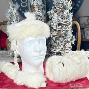 May include: A white faux fur hat, scarf, and muff set. The hat has ear flaps and a small peak. The scarf has a tail. The muff is rectangular. The set is displayed on a red surface.