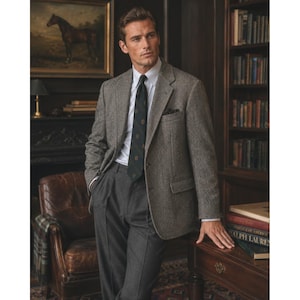 May include: A grey tweed blazer worn over a white collared shirt and a dark green patterned necktie, paired with charcoal grey trousers. The blazer features notched lapels, a chest pocket with a pocket square, and two front flap pockets.