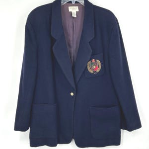 May include: Navy blue blazer with a gold button closure and two front pockets. The blazer features an embroidered crest on the chest with gold, red, and green details. The interior lining is a dark purple color.