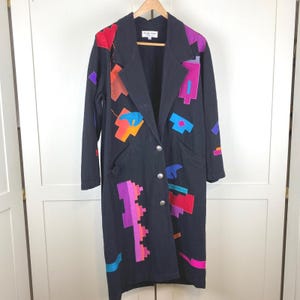 May include: A black long-line coat featuring a notched collar, three silver-toned buttons, and colorful geometric fabric appliqués in shades of red, orange, purple, pink, and blue. The interior label reads "i todo mas".