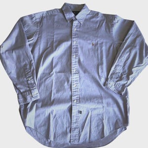 May include: A light blue button-down shirt with long sleeves. The shirt features a button-down collar, a small embroidered logo on the chest, and buttoned cuffs. The shirt is made of a soft, woven fabric.