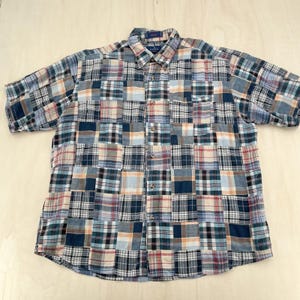 Può includere: Una camicia a maniche corte con bottoni caratterizzata da un design patchwork composto da vari quadrati di tessuto a quadri e scozzesi nelle tonalità blu navy, azzurro, bianco, beige e rosso tenue. La camicia include un colletto e una tasca.