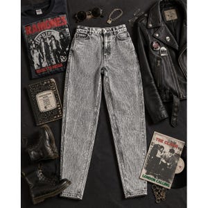 Peut inclure: Un jean gris délavé à taille haute entouré d'un t-shirt noir Ramones, d'une veste de moto en cuir noir avec des épinglettes, de bottines à lacets noires, d'un cahier de composition, de lunettes rondes, d'un collier chaîne et d'un disque The Clash.