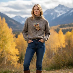 Könnte beinhalten: Ein melierter brauner Rollkragenpullover mit einer zentralen Stickerei eines Bären, der eine gelbe Weste und einen blauen Schal trägt, kombiniert mit einer dunkelblauen Jeans. Der Pullover hat lange Ärmel sowie gerippte Bündchen und Saum.