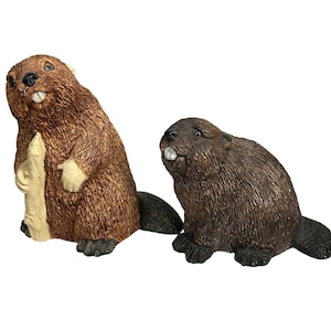 May include: Two detailed beaver figurines. One beaver is standing upright, holding a piece of wood, and the other is sitting. Both have brown fur, black tails, and white teeth. The figurines are likely made of resin or a similar material.