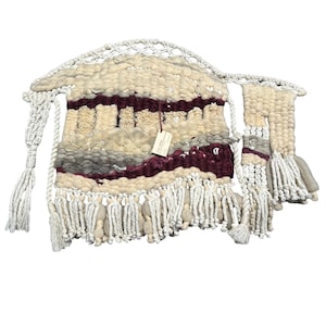 May include: A woven wall hanging with a cream, gray, and burgundy color scheme. The textile features a textured design with fringe and macrame details. A small tag hangs from the center of the piece. The wall hanging is a decorative home decor item.