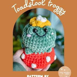 May include: A crocheted plush toy in the shape of a toadstool frog. The frog has a green body, a red base with white spots, and a yellow and white flower on its head. The text "Toadstool Froggy" and "Pattern by Cozy Season Crochet" are also visible.