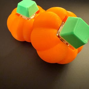 May include: Two orange, 3D-printed pumpkins with green square tops. The pumpkins have a ribbed texture and are set against a dark background. The green tops are square and appear to be attached to the pumpkins.