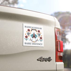 May include: A square sticker with the text "RADICALIZED BY BASIC DECENCY" and a floral design with a butterfly in the center. The sticker is on the back of a white vehicle.