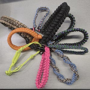 May include: A collection of colorful paracord wrist lanyards in various colors, including black, orange, pink, and blue. Each lanyard features a metal clasp for attaching to keys or other items. The lanyards are arranged in a circular pattern.
