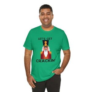 May include: A green t-shirt with a cartoon nutcracker design. The nutcracker wears a red coat and black hat, with the words "LET'S GET CRACKIN'" printed above and below the image in black font. The shirt is made of a soft material.