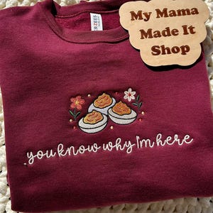 May include: Burgundy sweatshirt with embroidered deviled eggs and flowers. The text "you know why I'm here" is embroidered below the design. A wooden sign that says "My Mama Made It Shop" is on the top right.