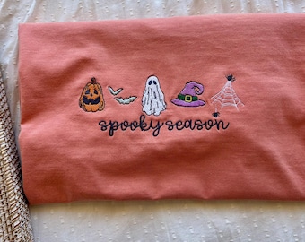 May include: Coral-colored t-shirt with embroidered Halloween-themed designs. Features a jack-o'-lantern, bats, a ghost, a witch's hat, and a spiderweb. The phrase "spooky season" is embroidered below the designs. The shirt is folded.