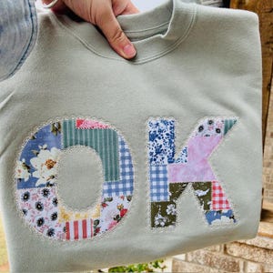 May include: Sage green sweatshirt with the letters "OK" appliqued on the front. The letters are made of patchwork fabric with floral, striped, and gingham patterns. The edges of the letters are trimmed with a scalloped lace.