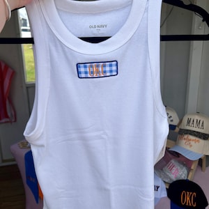 May include: White ribbed tank top with a blue and white checkered patch that reads "OKC" in orange lettering. The tank top is from Old Navy and is displayed on a black hanger.