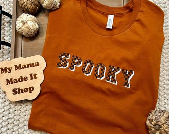 May include: An orange t-shirt with the word "SPOOKY" in a checkered pattern. The shirt is displayed on a wooden tray with decorative pumpkins. A wooden sign that says "My Mama Made It Shop" is also visible.