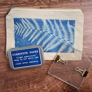 May include: A beige canvas zippered pouch featuring a blue fern leaf print, a rectangular metal tin labeled Cyanotype Paper with instructions, and a clear acrylic sheet with two gold-toned metal binder clips attached to the edges.