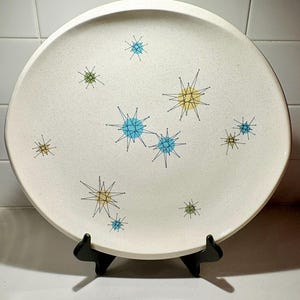 May include: A circular, off-white ceramic plate with a speckled finish. It features a design of stylized starbursts in yellow, blue, and green, outlined in black. The plate is presented on a black stand.