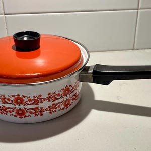 Puede incluir: Una cacerola vintage de esmalte blanco con tapa naranja y asa negra. La olla presenta un diseño floral en rojo. La tapa tiene un pomo negro. Esta batería de cocina retro es perfecta para cocinar.