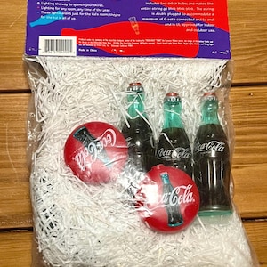 May include: Coca-Cola string light set with 10 lights and fun covers. The set includes two extra bulbs. The string is double plugged to accommodate a maximum of 6 sets. The packaging features the Coca-Cola logo and product information.