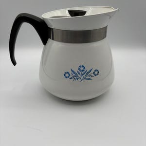 Könnte beinhalten: Weiße Vintage-Kaffeekanne mit schwarzem Griff und Deckel. Die Kanne hat ein silbernes Band und ein blaues Blumenmuster. Die Kanne, wahrscheinlich aus der Mitte des 20. Jahrhunderts, ist zum Brühen von Kaffee gedacht.