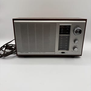 May include: A rectangular Realistic MTA-15 AM/FM table radio with a dark brown wood-grain finish cabinet and a silver front panel. The front features a horizontal speaker grille, a vertical frequency display, and three silver control knobs.