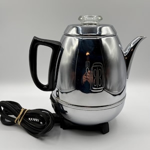 May include: A polished chrome electric percolator coffee pot featuring a black plastic handle, a clear glass knob on the lid, and a black power cord. The body of the coffee pot has a rounded shape with a spout and a black base with small feet.